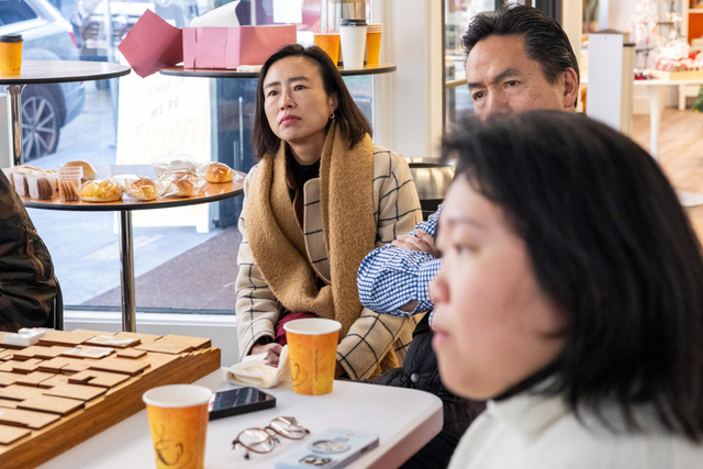 Three people sit at a table in a cafe, with two orange paper cups, pastries on a nearby table, and a wooden game or puzzle on the table.